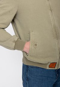 Persoon draagt een beige jas met geribbelde boorden en zoom, hand in de zijzak, blauwe spijkerbroek zichtbaar onder de jas.