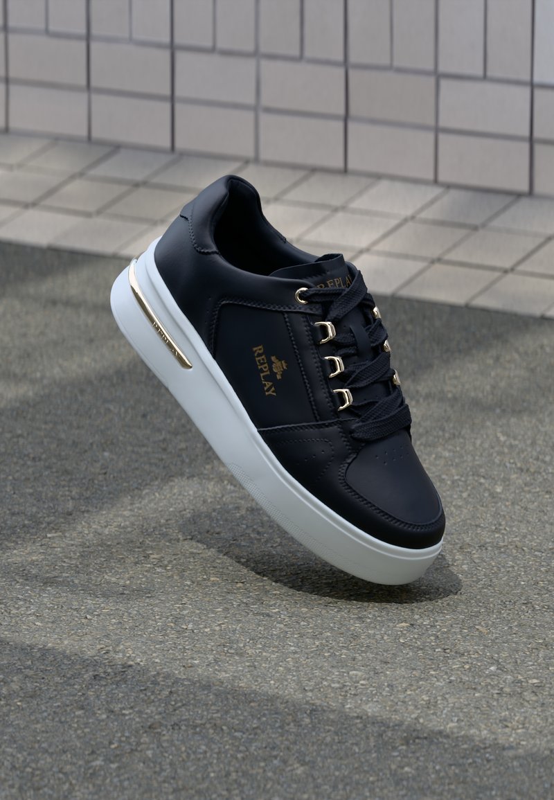 Navy leather sneaker with gold hardware, white sole, lace-up design, and "Replay" branding. Features perforations on the toe area.