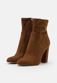 Par de botines de ante marrón con tacones altos gruesos y cremalleras laterales, mostrados sobre un fondo blanco liso.