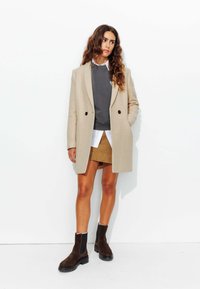 Manteau en mélange de laine beige, sweat gris, chemise blanche, jupe beige, bottines en daim marron, tissus texturés, design croisé, look superposé.