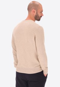 Uomo calvo con barba, che indossa un maglione beige e pantaloni neri, rivolto di lato con le mani in tasca su uno sfondo neutro.