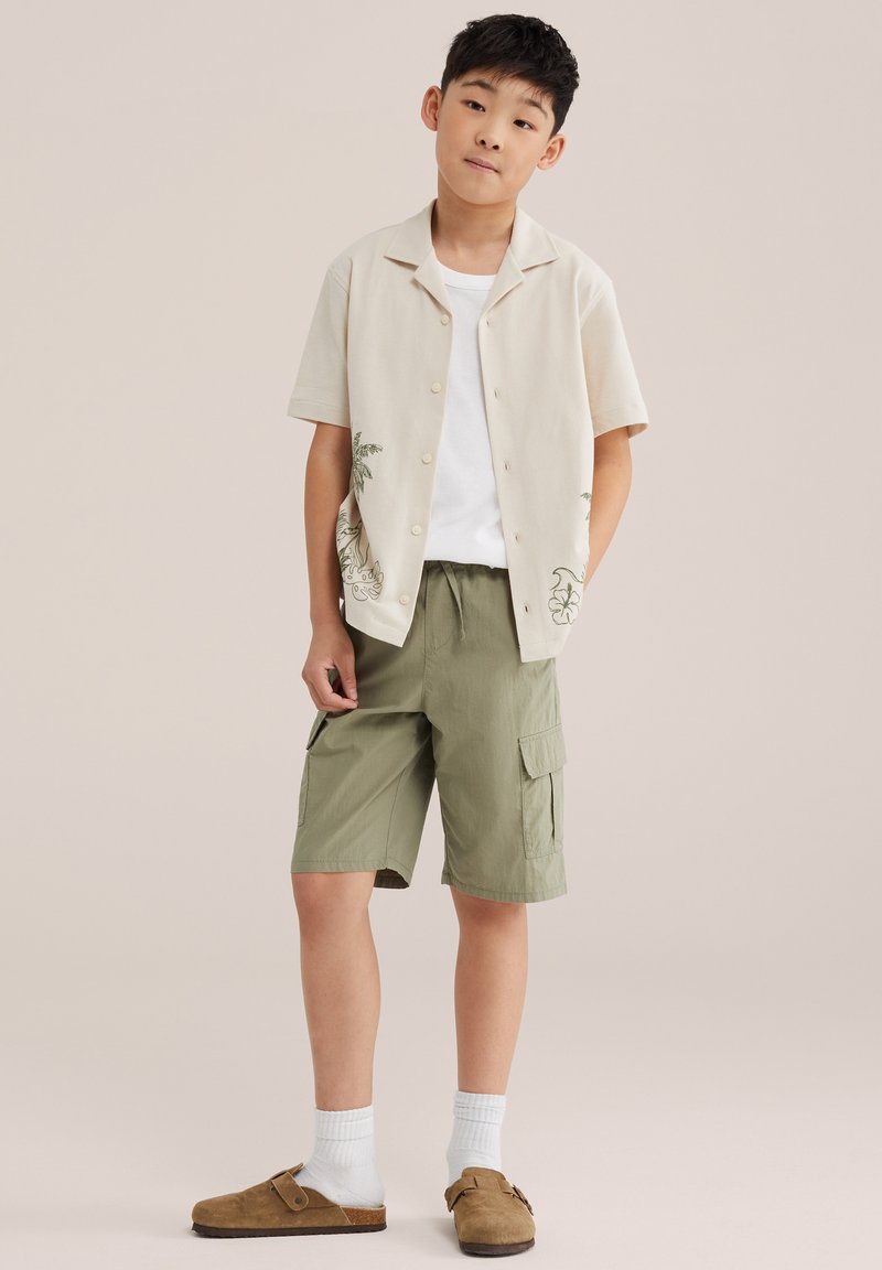 Garçon debout, les mains dans les poches des passants de ceinture, portant une chemise beige à manches courtes boutonnée, un t-shirt blanc, un short cargo olive, des chaussettes blanches et des chaussures marron sans lacets.