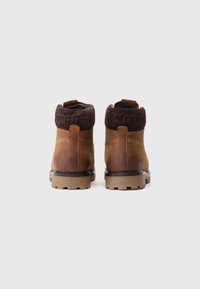 Bottines en cuir marron avec des cols rembourrés marron foncé et des semelles en caoutchouc épaisses, vues de l'arrière sur un fond uni.