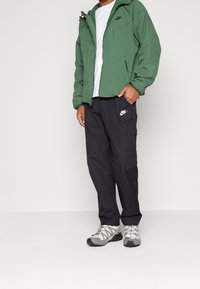 Grüne Zip-Jacke, weißes T-Shirt, schwarze Cargo-Hosen und graue Sneakers. Die Jacke hat elastische Bündchen; die Hosen verfügen über seitliche Taschen und ein Logo.