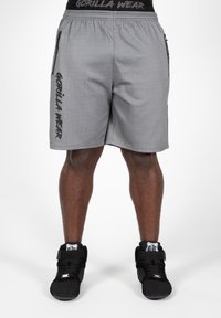Grijze sportbroekjes met een mesh-textuur, elastische tailleband, zijritsen en "GORILLA WEAR" gedrukt op het linkerpijpje. Zwarte hoge sneakers.
