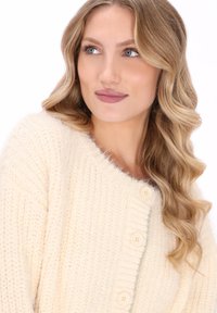 Blondynka o falowanych włosach, ubrana w kremowy sweter z dzianiny z zapięciem na górze, patrzy w prawo na tle białego tła.