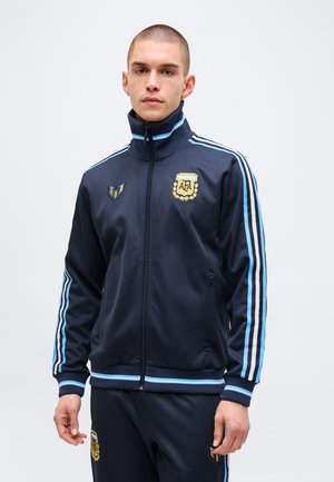 Jeune homme portant un survêtement bleu marine avec des rayures bleu clair et les logos de l'Association de football d'Argentine sur la veste et le pantalon.