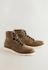Botas de tobillo de ante marrón con cordones en blanco y negro, punta redonda y suela de goma blanca. Cuenta con seis ojales metálicos para los cordones.