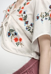 Μπεζ κοντομάνικο τοπ με κεντήματα λουλουδιών και δέσιμο μπροστά, φορεμένο πάνω από λευκό στρώμα και συνδυασμένο με μοβ παντελόνια.