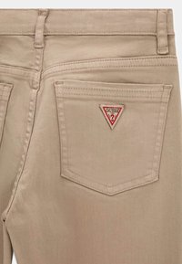 Beżowe spodnie o czystym designie, z dwoma tylnymi kieszeniami. Na prawej kieszeni znajduje się czerwona i biała trójkątna naszywka z logo "GUESS".