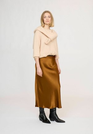 Femme aux cheveux blonds portant un pull beige, une jupe en satin marron et des bottines noires à talons, debout devant un fond clair uni.