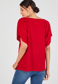 Donna con capelli scuri raccolti in uno chignon, che indossa una maglietta rossa corta e morbida con texture e jeans blu, voltata leggermente per mostrare il retro.