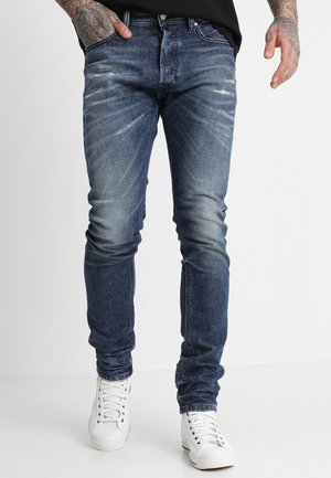 Mann trägt ausgewaschene, schmal geschnittene blaue Jeans mit leichten Used-Effekten, kombiniert mit weißen High-Top-Sneakers, steht mit einer Hand in der Tasche.