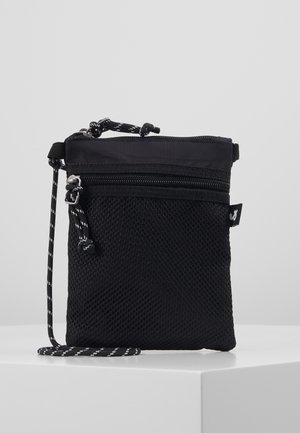 Mała czarna torba na ramię z siateczkową przednią kieszenią, zamknięciem na suwak i czarno-białym, regulowanym paskiem, eksponowana na białej powierzchni.