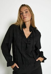 Blusa nera con scollo e polsini a balze. Realizzata in tessuto leggero con un delicato motivo a trama. Presenta una vestibilità comoda.