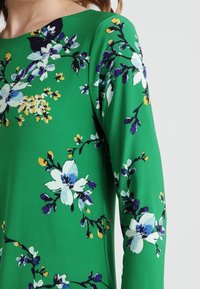 Zelené květinové šaty s bílými a modrými květinovými vzory a žlutými akcenty. Hladká textura, dlouhé rukávy a lodičkový v ýstřih.