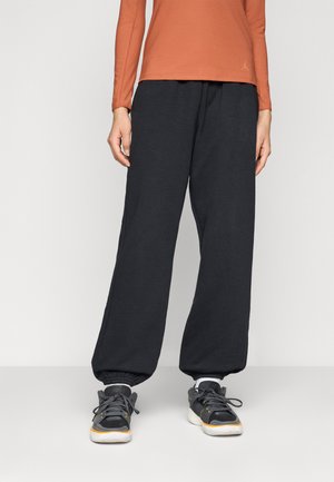 Person iført løse svarte sweatpants med elastiske mansjetter, svarte og grå joggesko med oransje detaljer, og en oransje langermet topp.