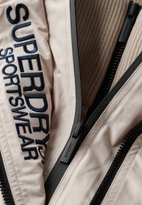 Lys beige sportsjakke med sorte lynlåse og syninger. Har broderet "SUPERDRY" logo i mørkeblå på stoffet med fin struktur.
