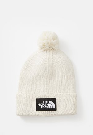 Beanie tricotat de culoare crem, cu o pom-pom în vârf și o etichetă rectangulară neagră pe manșetă, pe care este inscripționat "THE NORTH FACE".