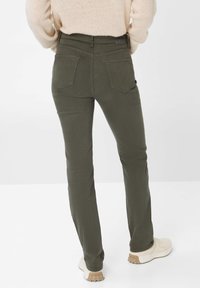 Olivgrüne Jeans mit enganliegender Passform, ausgestattet mit zwei aufgesetzten Taschen hinten und einem strukturierten Bund, kombiniert mit cremefarbenen Turnschuhen.