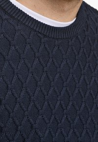 Marineblauer Strickpullover mit einem strukturierten Rautenmuster, ausgestattet mit einem gerippten Rundhalsausschnitt und einem darunter sichtbaren weißen Unterhemd.