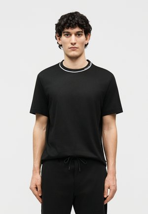 Ung mand med krøllet hår iført en sort kortærmet skjorte med hvid kant ved kraven og sorte bukser med snører.