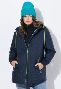 Giacca a vento blu marina trapuntata con zip frontale, dettagli verde neon e cappuccio. Abbinata a un berretto a coste turchese. Denim casual sotto.