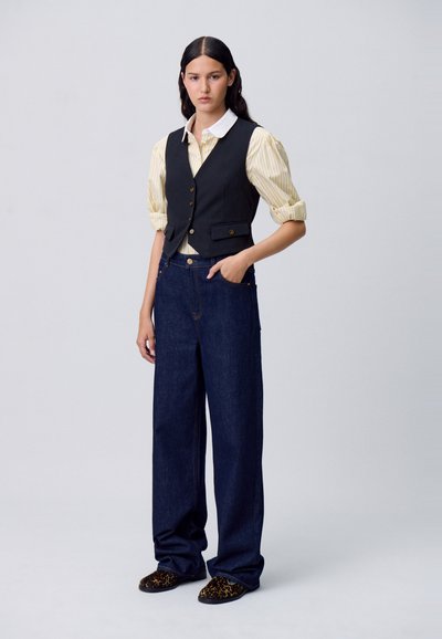 Una giovane donna sta in piedi con sicurezza, indossando un gilet nero sopra una camicia a righe e pantaloni di jeans larghi, abbinati a pantofole con motivo.