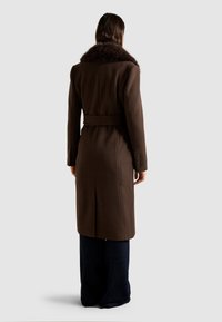 Abrigo de lana marrón con cuello de piel, cintura ceñida con cinturón y puños abotonados. Presenta un diseño largo y ajustado con una abertura en la parte trasera.