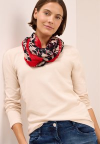 Écharpe à motifs rouge et marine, mêlant texte et formes, portée lâchement autour du cou, associée à un haut beige clair à manches longues.