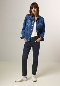 Jeansowa kurtka w ciemnoniebieskim kolorze z zapięciem na guziki, dwoma kieszeniami na piersi i luźnym fasonem. Uzupełniona o granatowe spodnie i białe sneakersy z zielonymi akcentami.