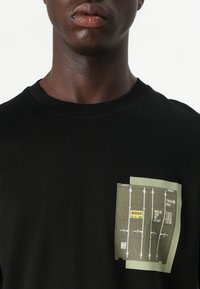 T-shirt nero a maniche corte con un design grafico rettangolare verde che presenta testo e illustrazioni a linee nella zona sinistra del petto.