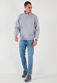 Suéter de pullover gris claro con cuello de media cremallera, costuras laterales y puños de canalé. Lleva puesto con vaqueros azules y zapatillas grises.
