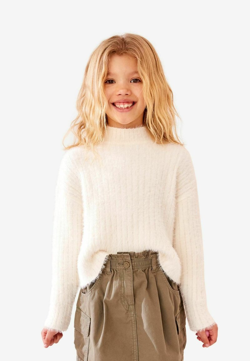 Weißer, gerippter Strickpullover mit hohem Kragen, kombiniert mit einem khakifarbenen Rock mit seitlichen Taschen und einem elastischen Bund.
