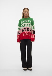 Groen en rood oversized sweater met de tekst "SANTA PLEASE STOP HERE" in verschillende kleuren. Zwarte wijdvallende broek. Effen achtergrond.