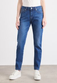 Persona che indossa jeans blu a vita media e sneakers bianche, in piedi su un pavimento semplice contro uno sfondo bianco.