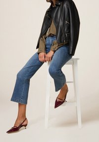 Chaqueta de motociclista de cuero negro, camisa oliva, jeans cropped azules con dobladillo deshilachado y tacones slingback de charol burdeos. Sentada en un taburete blanco.