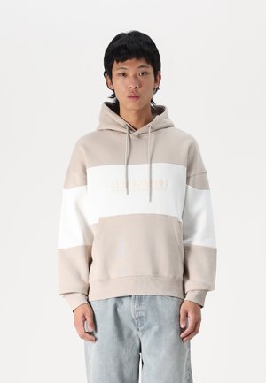 Sweatshirt à capuche en blocs beige et blanc, avec un tissu texturé, une poche kangourou et un logo de marque brodé sur la poitrine.