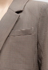 Καφέ και μπεζ σακάκι με μοτίβο houndstooth, γιακά με πέτο και τσέπη στήθους, φορεμένο στο πάνω μέρος του κορμού.