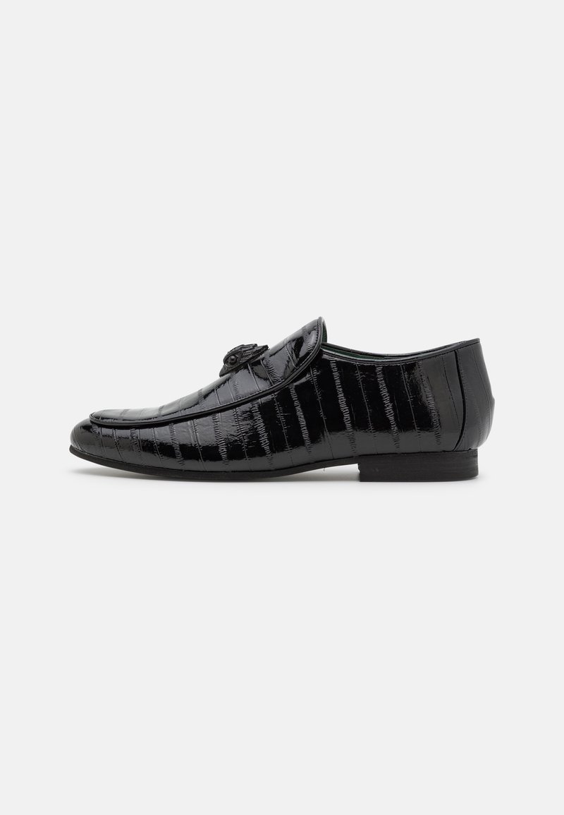 Mocassin en cuir noir brillant avec texture crocodile et pompon décoratif sur le dessus, talon plat, vue de profil sur fond blanc.