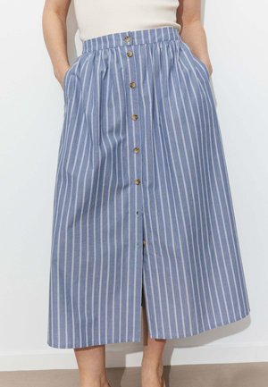 Femme portant une jupe midi à rayures verticales bleues et blanches avec des boutons marron à l'avant et les mains dans les poches, associée à un haut blanc sans manches.