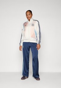 Balta striukė su pasteliniais rožiniais ir mėlynais akcentais, su juodomis juostelėmis. Tamsiai mėlynos treniruočių kelnės su šviesiai mėlyna juostele ir logotipu.