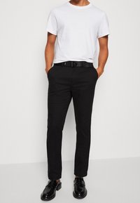 Schwarze, schmal geschnittene Hose mit Seitentaschen und Gürtel. Kombiniert mit einem schlichten weißen T-Shirt mit rundem Ausschnitt und schwarzen Loafers.