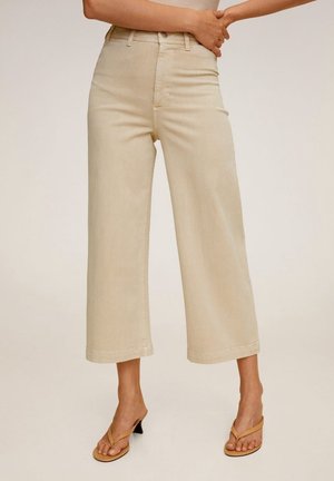 Kobieta w beżowych spodniach z wysokim stanem i szerokimi nogawkami oraz w brązowych sandałach na obcasie, stojąca z założonymi na siebie rękami na wysokości talii.