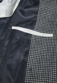 Graues Hahnentritt-Gewebe auf der Außenseite, marineblaue Satin-Futter mit einer kontrastierenden gestreiften Tasche, die ein strukturiertes Design und Textur präsentiert.