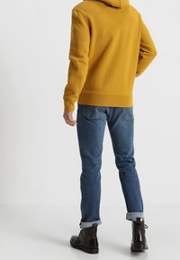 Felpa gialla senape con finitura testurizzata, dotata di cappuccio posteriore e polsini a coste, abbinata a jeans blu e stivali neri alla caviglia.