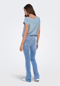 Γυναίκα που φοράει ανοιχτό μπλε κοντομάνικο τοπ με φερμουάρ στην πλάτη, ανοιχτό μπλε τζιν παντελόνι με ίσια γραμμή και λευκά παπούτσια, όρθια με την πλάτη στραμμένη.