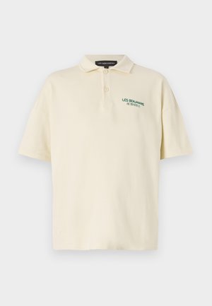 Kerrat polo vaalealla värillä vihreällä tekstillä, vaaleat housut logokuvioilla, musta laukku pidettynä yhdellä kädellä, sekä mustat vetoketjulliset saappaat valkoisten sukkien kanssa.