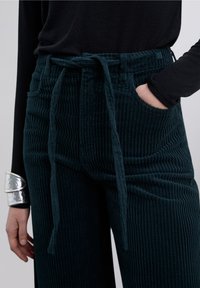 Pantalon en velours côtelé vert sarcelle foncé avec nervures verticales, taille à cordon de serrage et poches avant. La texture semble douce et légèrement moelleuse.