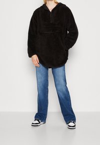 Persona che indossa un pullover nero peloso con tasca frontale, jeans blu e sneakers bianco e nere, in piedi contro uno sfondo bianco.